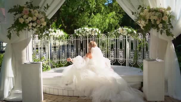 Noiva em vestido frágil sentado em rotunda decorado com flores e cortinas — Vídeo de Stock