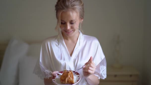 Bela mulher em robe de seda comer bolo no quarto. manhã antes do casamento . — Vídeo de Stock