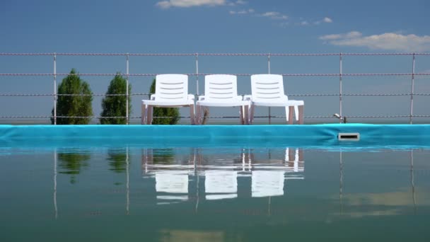 Tumbonas en el cielo azul de pie cerca de la piscina y se reflejan en el agua — Vídeos de Stock