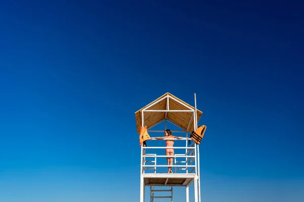 stock image rear view. teenage girl stands on lifeguard tower on beach against cloudless sky.