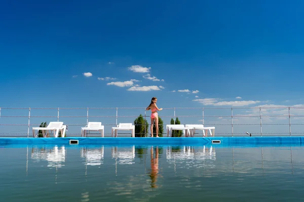 Vista trasera. chica en traje de baño se encuentra entre tumbonas por el cielo de fondo de la piscina —  Fotos de Stock