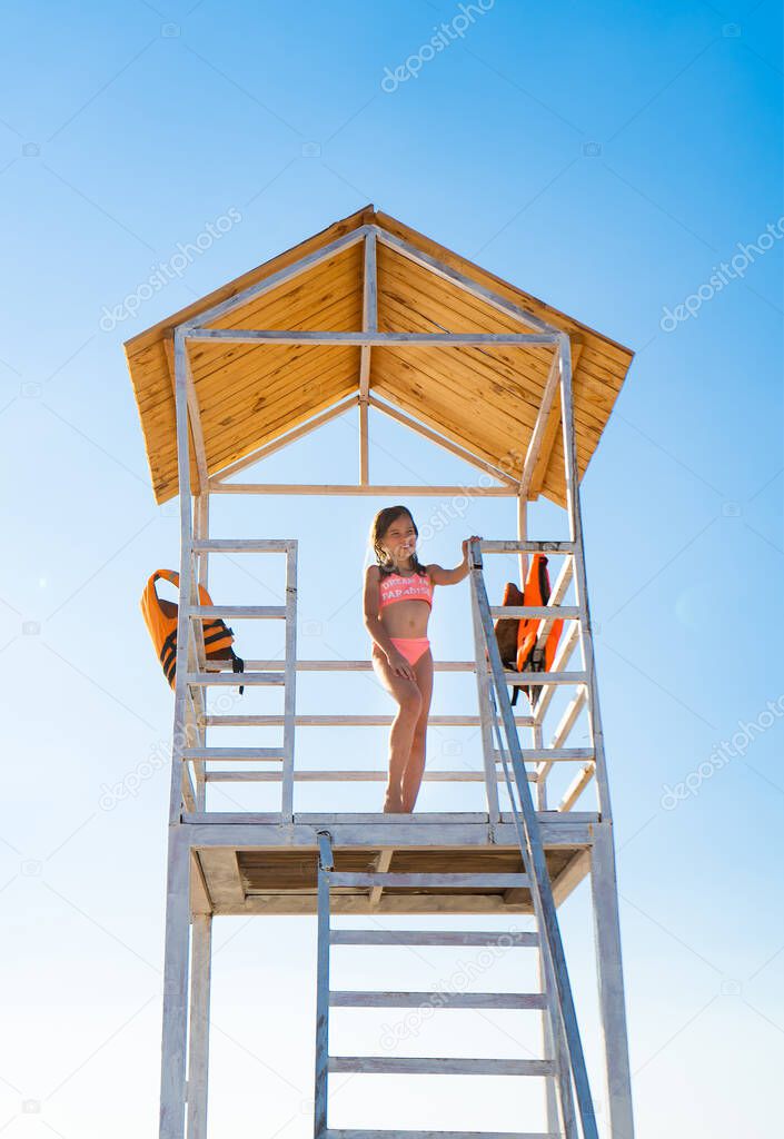 teenage girl stands on lifeguard tower on beach against cloudless sky.