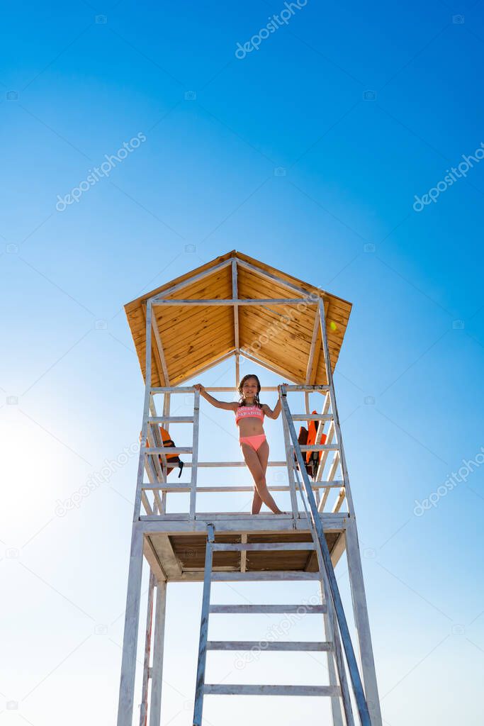 teenage girl stands on lifeguard tower on beach against cloudless sky.