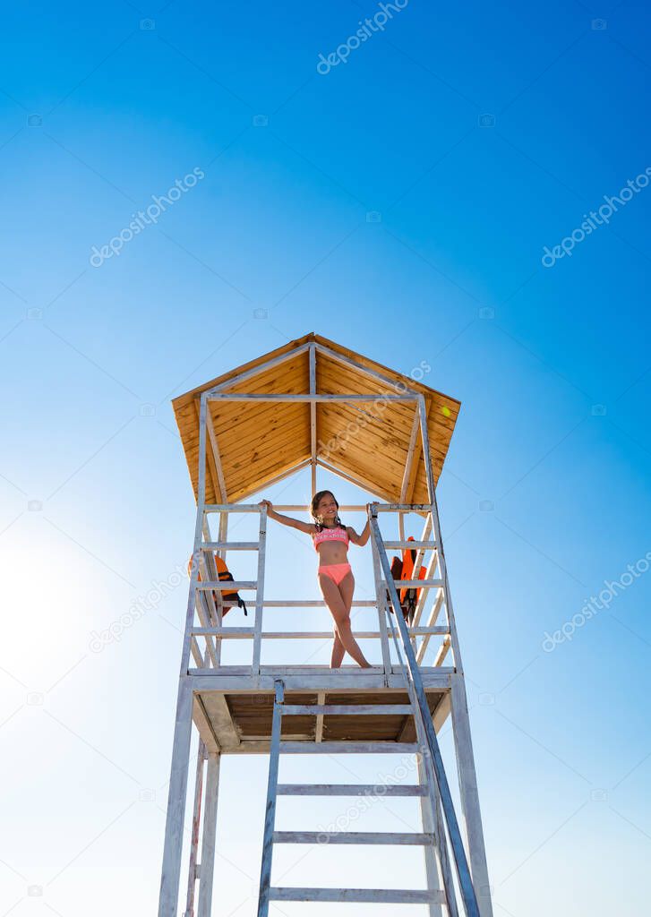 teenage girl stands on lifeguard tower on beach against cloudless sky.