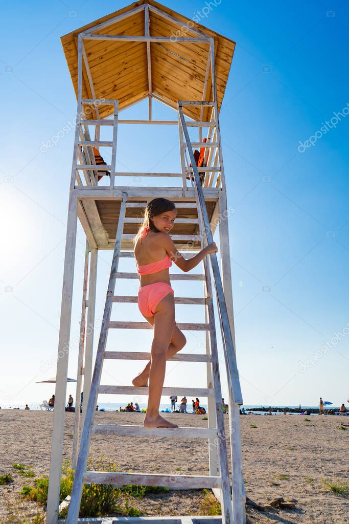 teenage girl goes up stairs lifeguard tower on beach against cloudless sky.