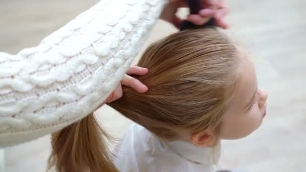 Vista lateral. dulce escuela chica bosteza mientras madre peina cabello y recoge en la cola — Vídeo de stock