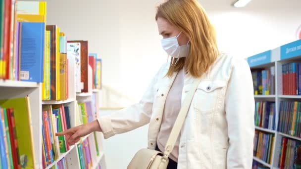 Femme dans un masque médical de protection choisit un livre dans un magasin ou une bibliothèque. — Video