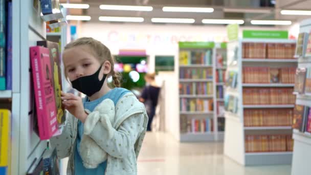 保護医療マスクの子供の女の子は店や図書館の棚に本を選ぶ。ロストフ。ロシアだ。2020年9月29日 — ストック動画