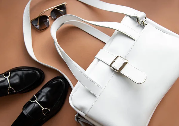 Woman casual fashion clothes and accessory,  top view on brown background. White leather bag, black shoes loafer, sunglasses , flat lay.