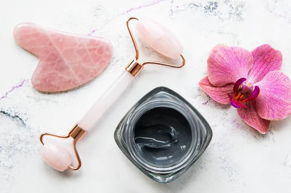 Ev banyosunda cilt bakımı rutini - mermer arka planda sağlık kozmetik terapisi, maske ve orkide çiçekleri için yeşim taşlı masaj aleti