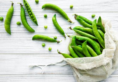 Beyaz ahşap arka planda taze bezelye kabuklarıyla dolu yeniden kullanılabilir çevre dostu çanta.