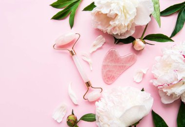 Yüz masajı terapisi, masaj yağları ve pembe şakayıklar için yeşim bir yüz silindiri. Pembe pastel arka planda düz uzandı