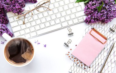 Üst kattaki ofis masasında yatıyordu. Klavyeli çalışma alanı, leylak çiçekleri ve beyaz arka planda ofis malzemeleri.