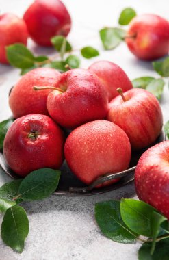 Olgun kırmızı elmalar ve su damlacıkları, metal bir kapta otururlar, yeşil yapraklar eşliğinde desenli bir yüzeyde.