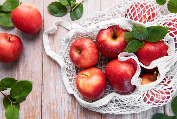 Yapraklı olgun kırmızı elmalar, yeniden kullanılabilir bir alışveriş çantasının içinde, tahta bir masada, sürdürülebilir bir yaşam tarzını sembolize ediyor.