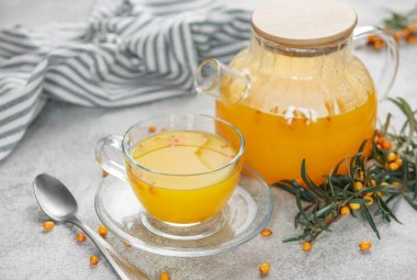 Cam çaydanlık ve fincanda deniz dikenli çay, böğürtlen ve gri beton arka planda bir dal.