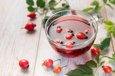 Taze demlenmiş gül çayı, gül kalçası ve yapraklarla çevrili, kırsal ahşap bir masada cam bir fincanda buharlaşarak oturur.