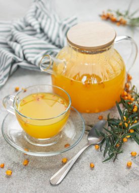 Cam çaydanlık ve fincanda deniz dikenli çay, böğürtlen ve gri beton arka planda bir dal.