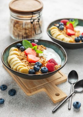 İçinde yoğurt, tahıl gevreği, muz dilimleri, çilek, ahududu ve yabanmersini bulunan iki kase sağlıklı kahvaltı.