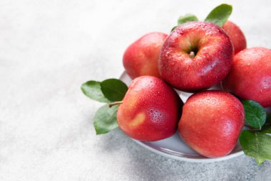 Su damlaları ve yeşil yapraklı olgun kırmızı elmalar, beyaz bir tabakta sıralanmış, taze ve sağlıklı bir görüntü yaratıyor.