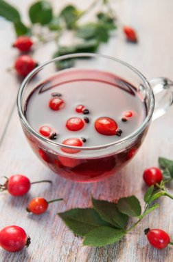 Taze demlenmiş gül çayı, gül kalçası ve yapraklarla çevrili, kırsal ahşap bir masada cam bir fincanda buharlaşarak oturur.