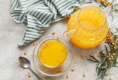 Cam çaydanlık ve fincanda deniz dikenli çay, böğürtlen ve gri beton arka planda bir dal.