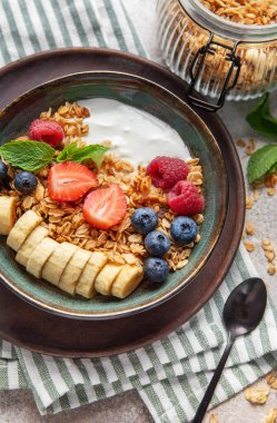 İçinde yoğurt, tahıl gevreği, muz dilimleri, çilek, yabanmersini, ahududu ve nane yaprakları bulunan bir kase sağlıklı kahvaltı.