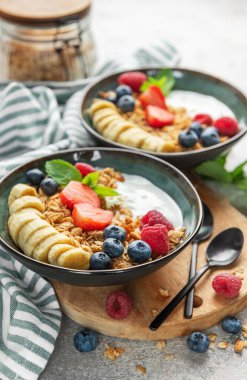 İçinde yoğurt, granola, muz dilimleri, çilek, ahududu ve yabanmersini bulunan iki kase sağlıklı kahvaltı yenmeye hazır.