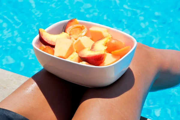 Tanned beautiful legs. Young woman relaxing and eating fruits. Fruits in a bowl by the pool near a thin slender fit girl legs. Fruit salad, healthy food. Summer concept