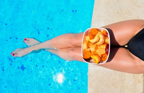 Krásné opálené nohy. Mladá žena uvolňující a jíst ovoce. Ovoce v míse u bazénu v blízkosti tenký štíhlý fit dívka nohy. Ovocný salát, zdravé jídlo. Letní koncept. — Stock fotografie