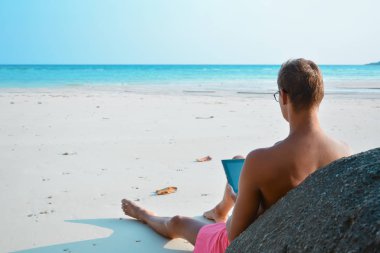 Kumsalda dizüstü bilgisayarıyla çalışan genç adam. Deniz kıyısında dizüstü bilgisayarı olan genç bir adamın arkasından görüntü. Sahilde serbest çalışan.