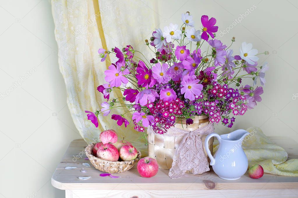 Still life with a bouquet of pink flowers in a vase and a basket of natural, ripe apples on a light background