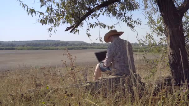 Чоловік працює поза офісом з ноутбуком, сидячи на березі озера — стокове відео