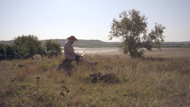 Hombre trabajando fuera de la oficina con el ordenador portátil sentado en la orilla del lago — Vídeos de Stock