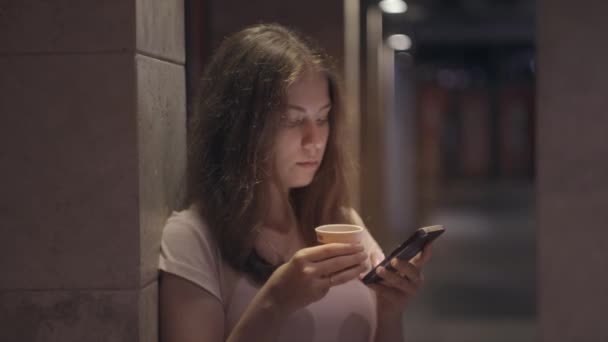 Retrato de una chica con café y un smartphone en una ciudad nocturna — Vídeos de Stock