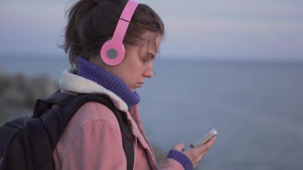 Close Up retrato de uma jovem mulher com rosto natural em pé na praia com um smartphone ao pôr do sol — Vídeo de Stock