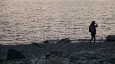 Gün batımında sahilde akıllı telefon ve kulaklıkla selfie çeken genç bir kadın.