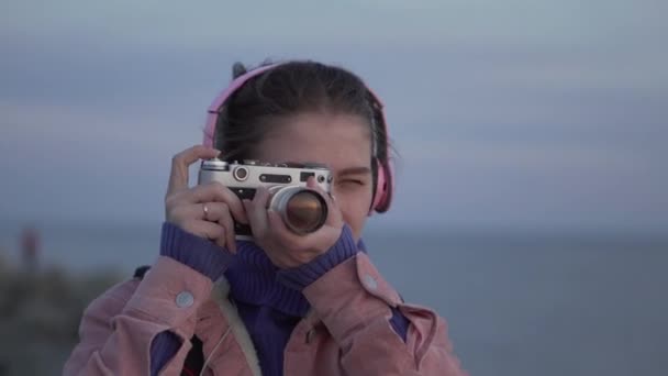 Menina em fones de ouvido tira uma foto do pôr do sol do mar em sua câmera vintage — Vídeo de Stock