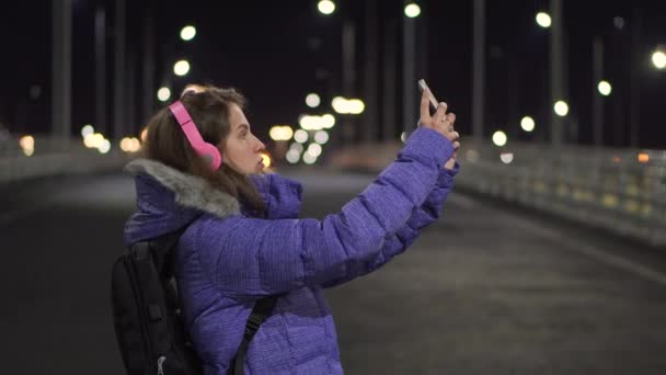 夜の町の街を歩く間に携帯電話を使ってビデオ通話で話す少女 — ストック動画