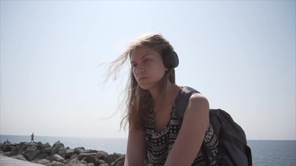 Una chica con una mochila y auriculares se sienta en el paseo marítimo. Movimiento lento — Vídeos de Stock