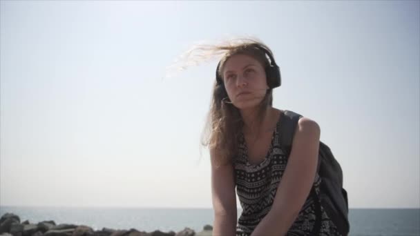 Una chica con una mochila y auriculares se sienta en el paseo marítimo. Movimiento lento — Vídeos de Stock