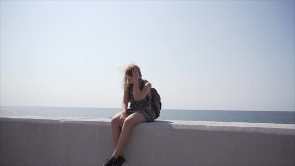 Una chica con una mochila y auriculares se sienta en el paseo marítimo. Movimiento lento — Vídeos de Stock