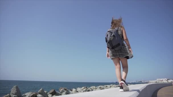 Flicka med en ryggsäck och hörlurar lyssnar på musik. Promenader längs strandpromenaden. — Stockvideo