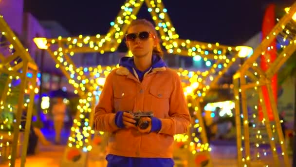 Uma menina de óculos com uma câmera de filme está contra uma estrela brilhante em um ambiente urbano — Vídeo de Stock