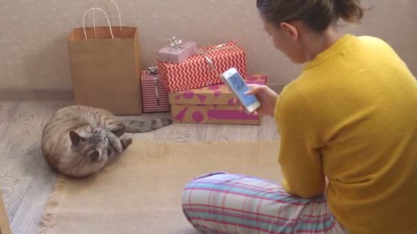 Kot bawi się świąteczną zabawką na podłodze w pokoju. Dziewczyna zdejmuje go z telefonu.. — Wideo stockowe