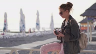 Güneş gözlüklü ve kot ceketli bir kız. Plajda film kamerası var. Gün batımının fotoğraflarını çekiyor..