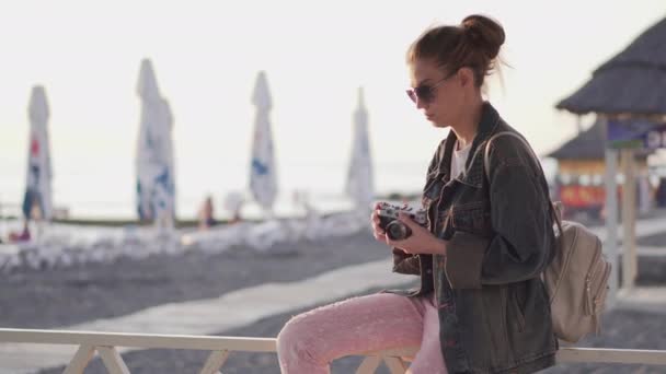 戴着太阳镜 身穿斜纹棉布夹克的女孩带着胶卷相机在海滩上拍摄海落时分的照片 — 图库视频影像