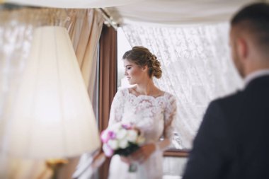 güzel ve zarif sarışın gelin uzun beyaz bir elbise ile mavi takımı ayakta pencere yakınındaki otel odasında ona yakışıklı gelin