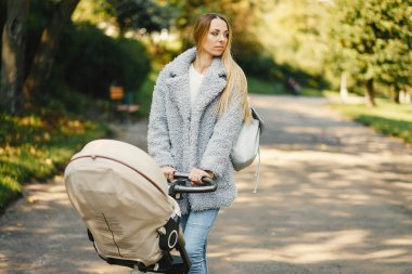 şık genç sarışın anne çevresinde güneşli Park içinde bebek ile bir bebek arabası itme