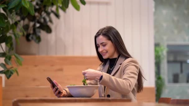 Видео, где девушка ест салат и пользуется мобильным телефоном — стоковое видео